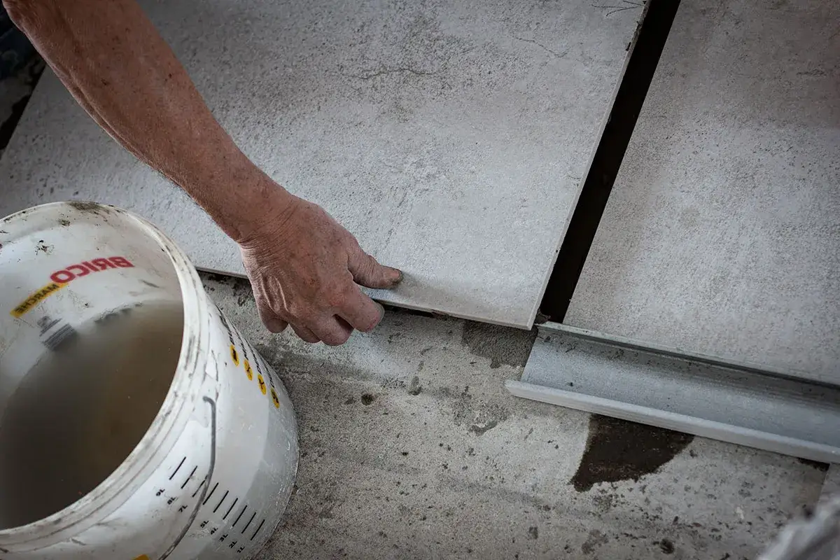 Dłoń kładzie płytkę ceramiczną obok wiadra z wodą.